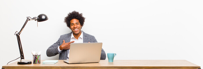 young black businessman looking happy, surprised, proud and excited, pointing to self on a desk