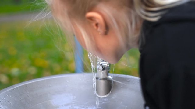 Cute adorable caucasian litte blond girl drinking water from faucet with drinking water fountain at city park near children playground at autumn outdoors. Drinkable water public supply for people