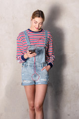 young blond girl standing by wall with mobile phone and listening to music with earphones © mimagephotos