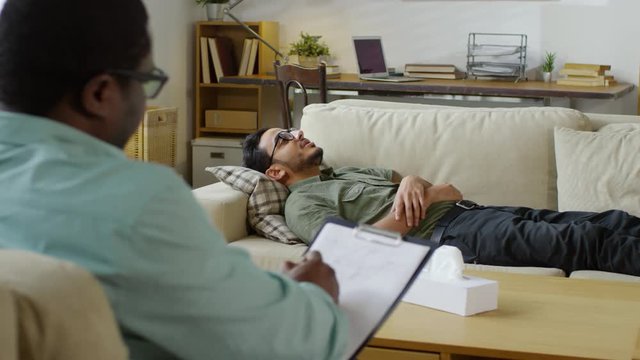 Young middle eastern man lying on sofa and telling about problems to african american psychotherapist during counseling session