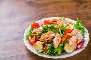 Fresh salad with fish, arugula, eggs,red pepper, lettuce, fresh sald leaves and tomato on a white plate on wooden table background