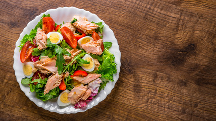 Fresh salad with fish, arugula, eggs,red pepper, lettuce, fresh sald leaves and tomato on a white plate on wooden table background