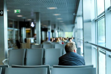 Back view of man sitting in waiting room at airport, wearing grey suit and glasses.