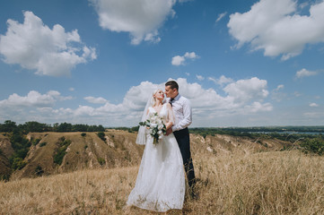 Beautiful newlyweds stand against the backdrop of nature, sky and clouds. Stylish groom in a white shirt hugs a smiling blonde bride. Concept and photography.