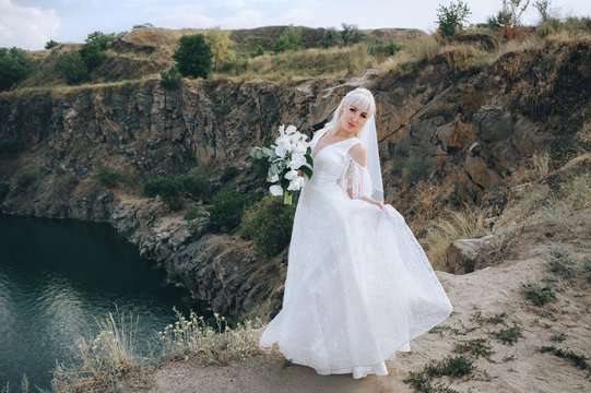 Beautiful blonde bride stands and dances against the backdrop of rocks, cliffs on a stone, holding on to a dress. Wedding portrait of a cute and smiling girl. Photography and concept.