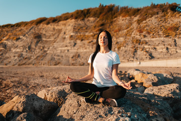 Naklejka na ściany i meble The concept of yoga and outdoor sports. Young woman doing yoga sitting on a large stone. In the background is a mountain