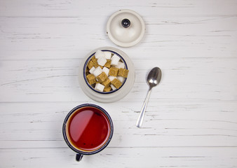 tea in a white enameled cup, cane sugar and white cubed sugar cubes in a sugar bowl tea spoon on a light wooden background