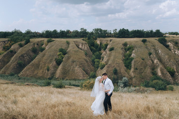 Fototapeta premium Stylish groom in a white shirt hugs a beautiful and smiling blonde bride against the background of nature, hills. Wedding portrait of lovers newlyweds. Photography and concept.