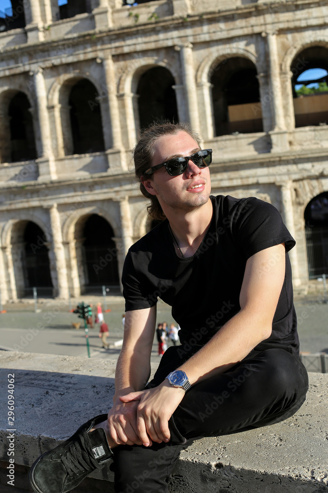 Wall mural young man sitting near colosseum background in rome, italy.