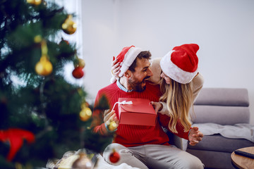 Excited handsome bearded caucasian man receiving gift from his girlfriend. Both having santa hats on heads. In foreground is christmas tree. Living room interior.