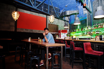 Portrait of handsome successful bearded south asian, young indian freelancer in blue jeans shirt sitting in night club against bar counter with cocktail and having a rest.
