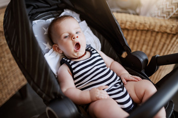 Sleepy adorable Caucasian 6 months old baby boy sitting in stroller in cafe and yawning.
