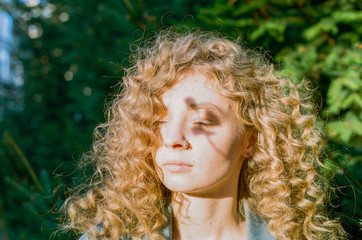 A young adult beautiful girl with blond curly hair stands on a green background with her eyes...