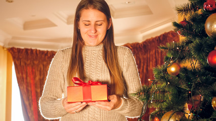 Toned portrait of happy smiling young woman standing with gift box next to Christmas tree in living room