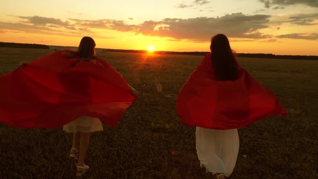children and mom play superheroes running across the field in the sun. Mom and daughter play super heroes in red cloaks at sunset in park. woman, savior, winner
