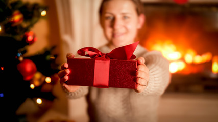 Fototapeta na wymiar Closeup image of young woman showing red Christmas gift box in camera while sitting next to Xmas tree and burning fireplace