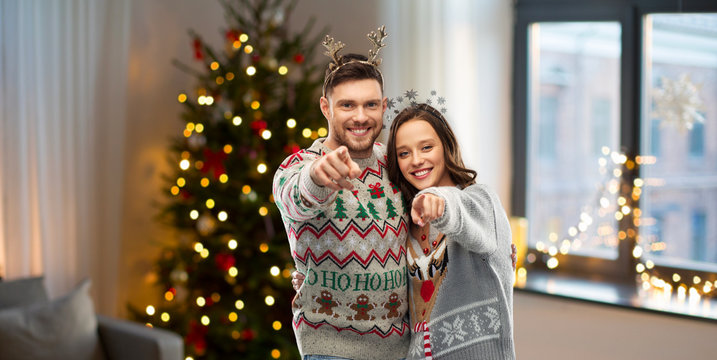 christmas, people and holidays concept - happy couple at ugly sweater party pointing at you over home background
