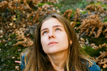 A beautiful young adult girl with blond hair and blue eyes sits on green autumn grass and moss against the background of dry yellow autumn leaves and looks up at the sky