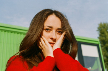 Brunette with a long hair in a red sweater holds her hands on her face, stands against the sky