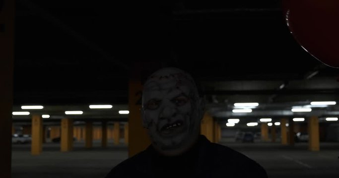 Maniac in leather mask is walking in dark parking lot, he&rsquo;s slowly walking straight at camera, he has red ball in his hand, scares passers-by, portrait shooting, monster from movie, Halloween costume