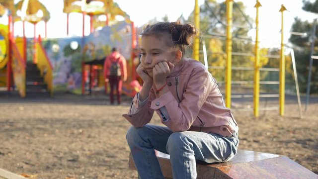 Sad lonely girl sitting on the background of a bright children's Playground where they play and have fun other children.