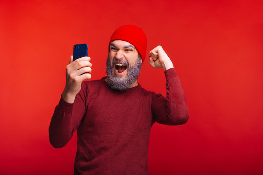 Photo of cheerful man with white beard and red cap looking at smartphone and celebrating success