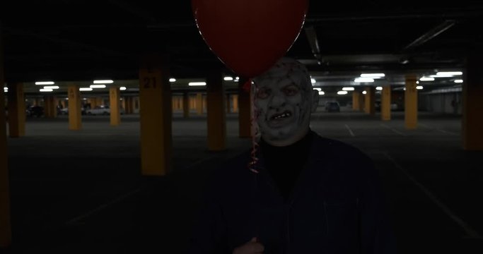 Scary man in creepy leather mask is standing with red ball in dark parking lot, maniac costume, dangerous man, scaring people for Halloween, creepy image, pretending to be dead zombie, alien from hell