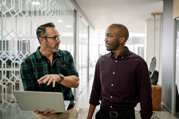 Two businessmen walking to boardroom for meeting discussing information on laptop