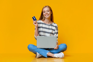 Cheerful girl holding credit card and laptop, sitting on floor - obrazy, fototapety, plakaty