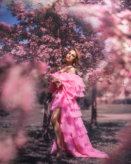 Art fashion portrait of a beautiful young blonde woman in a spring pink blooming garden in a long pink dress like a princess in fairytale