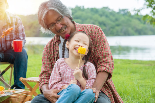 Little Asian Girl Enjoy With Eating Grilled Corn With Her Grandfather At Camping Site.