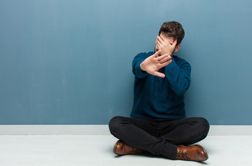 young handsome man sitting on floor covering face with hand and putting other hand up front to stop...