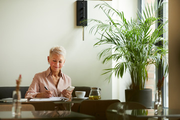 Mature smiling businesswoman using her mobile phone and making notes at the table in cafe