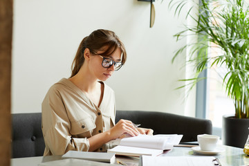 Serious young businesswoman in eyeglasses sitting at her workplace and looking notes in her notepad