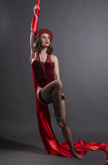 Beautiful girl wearing red beret and body and stockings elegantly posing in burlesque style, holding on to the fabric hanging from above. Fashionable, advertising, art design.