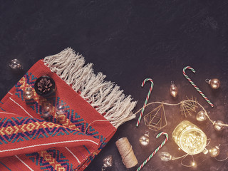 Christmas composition. A red knitted fringed plaid is on a stone background. Nearby are various Christmas decorations (balls, candy, red berries, a luminous garland in a glass jar). Flat lay.