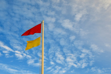 Top of a pole with a red and a yellow warning flags, indicating total absence of surveillance on the beach, against a cloudy blue sky, Italy