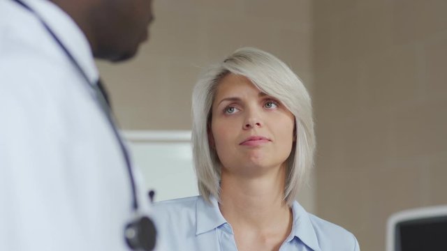 Closeup tracking shot of blonde female patient talking to unrecognizable African American general practitioner in white coat