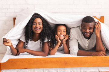 Cheerful black family of three hiding under blanket on bed