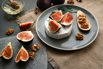 Figs slices on a round of camembert cheese on a gray plate with a leaf of fig tree and walnuts nearby. On a linen napkin. honey with wooden spoon in the background. Horizontal orientation.