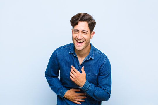 young handsome man laughing out loud at some hilarious joke, feeling happy and cheerful, having fun against blue background