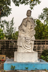 Fototapeta premium Bang Saen, Thailand - March 16, 2019: Wang Saensuk Buddhist Monastery. White stone Christian statue of Holy Family, Joseph, Mary and boy Jesus against silver sky and green foliage.