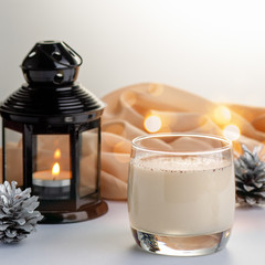 Traditional Christmas cocktail Eggnog with eggs, alcohol, grated nutmeg and cinnamon closeup. Sweet traditional drink on grey table with beige decorations, burning lanterns and pine cones