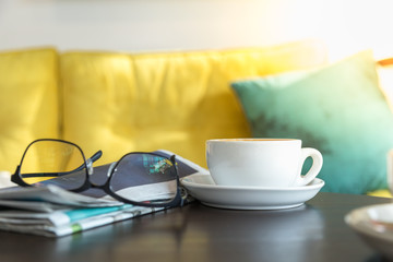 Close up of white cup of hot coffee latte with newspaper and reading glasses on wooden table in restaurant.