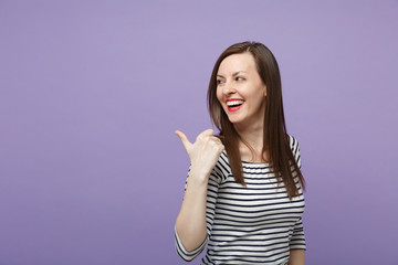Laughing cheerful young brunette woman girl in casual striped clothes posing isolated on violet purple background studio portrait. People lifestyle concept. Mock up copy space. Pointing thumb aside.