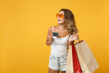 Cheerful young woman girl in eyeglasses posing isolated on yellow orange background. People lifestyle concept. Mock up copy space. Holding package bag with purchases after shopping, credit bank card.