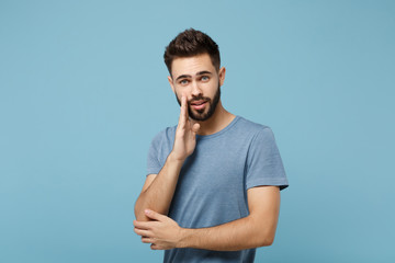 Young attractive man in casual clothes posing isolated on blue background in studio. People sincere emotions lifestyle concept. Mock up copy space. Whispers gossip and tells secret with hand gesture.