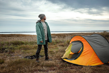 A bearded man near a camping tent in orange on the background of nature and the lake. The concept...