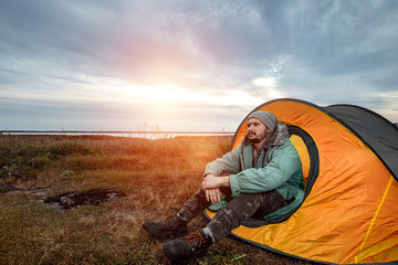 A bearded man sits camping in a tent against the backdrop of nature and the lake. The concept of travel, tourism, camping.