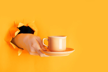 Hand holding a cup of coffee through a hole in torn saffron or light orange paper wall. Coffee...